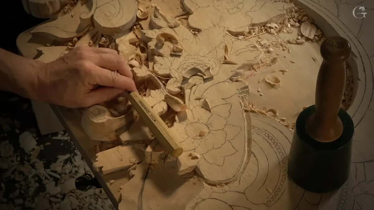 Overhead, one hand steers a slim tool with a pale handle into the carved scroll