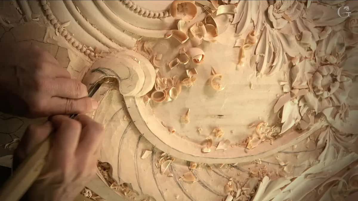Both hands work a gouge along the edge of the scroll, pencil lines still showing on the wood around it