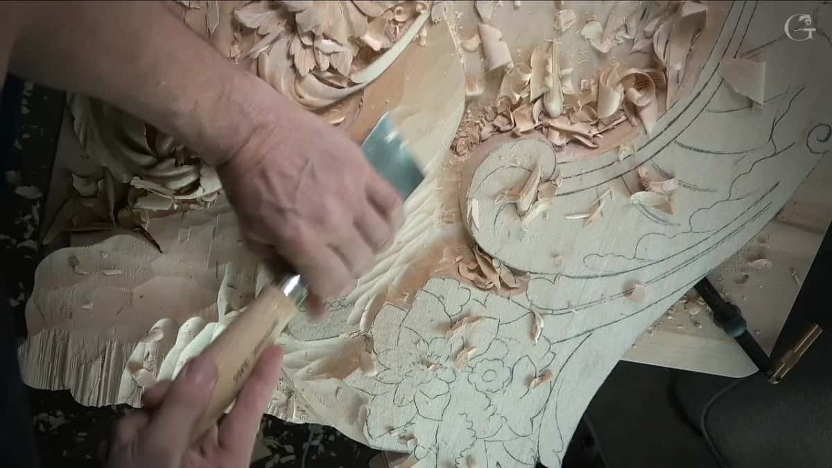 Both hands steering a wide, nearly flat gouge across the slope between the scroll and the flower drawing
