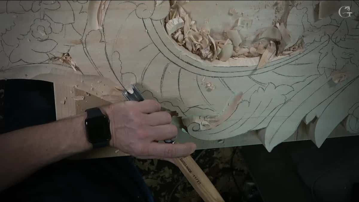 Fingers wrapped round a narrow gouge at the corner of the board, shavings behind it
