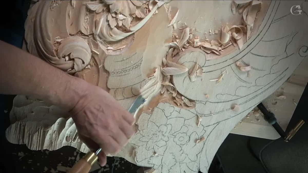 Wide gouge scooping beside a carved scroll, curled chips lying on the drawn flower