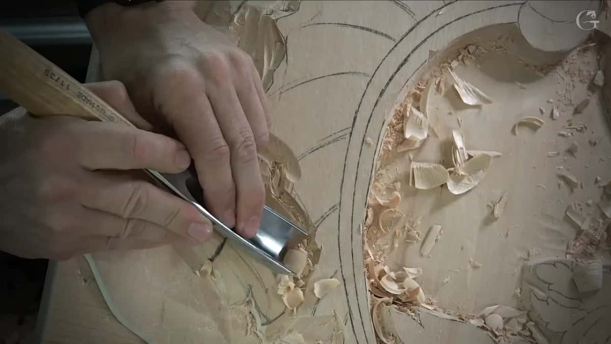 Two hands drive a wide U shaped gouge into the long carved shape beside the oval