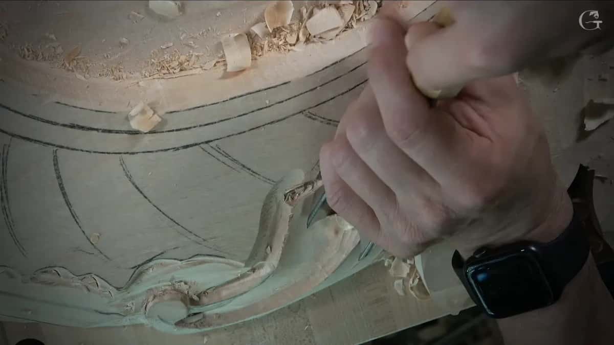 Low side view, a hand holds a gouge against the carving near the pencil bands