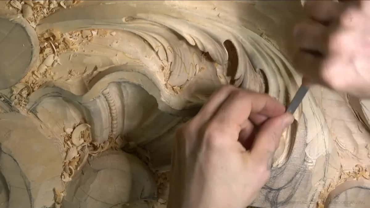 Close over the leaf edge, his thumb and finger pinch a short grey rasp while shavings lie scattered across the panel.