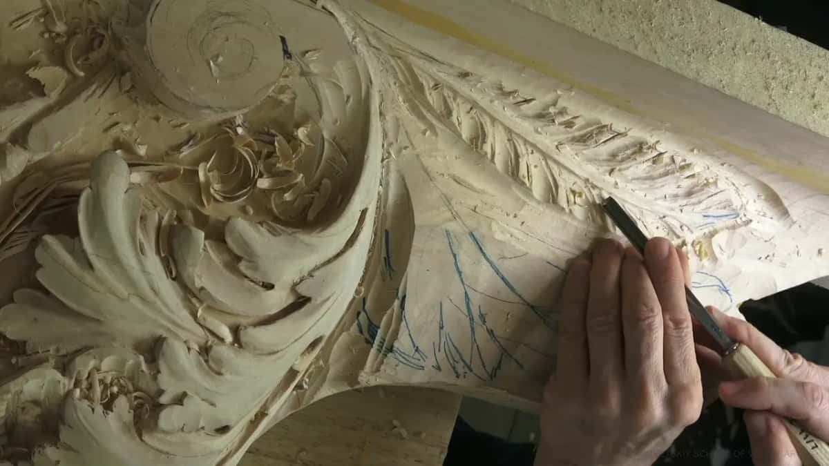 Both hands keep a long straight gouge low against the lower leaf, just under the row of carved ribs.