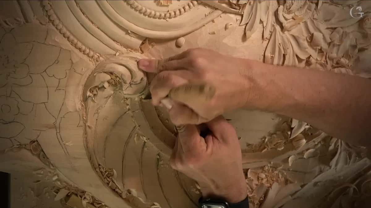 Seen from the far side of the bench, one hand drives a pale handled gouge into the curl while the other guides the blade