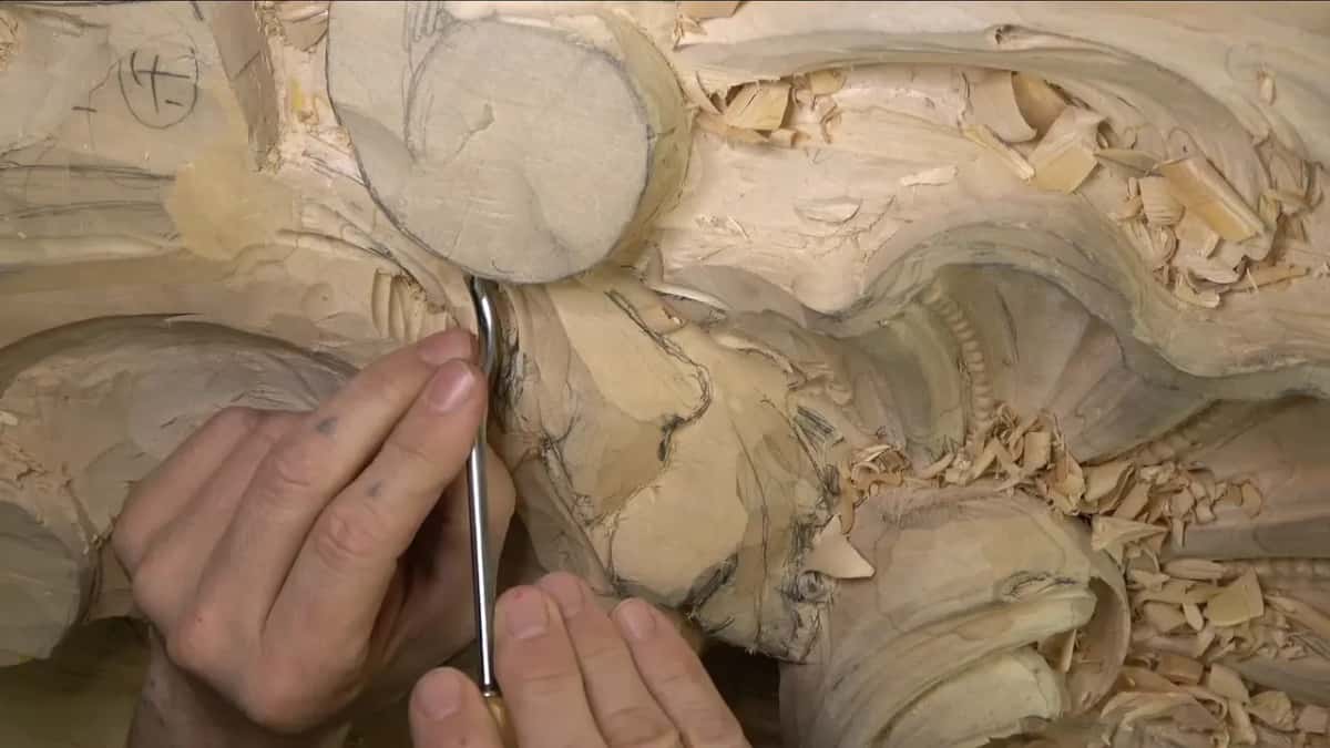 Fingertips steer a slim straight gouge down into the wood just below the round boss with pencil lines on it.