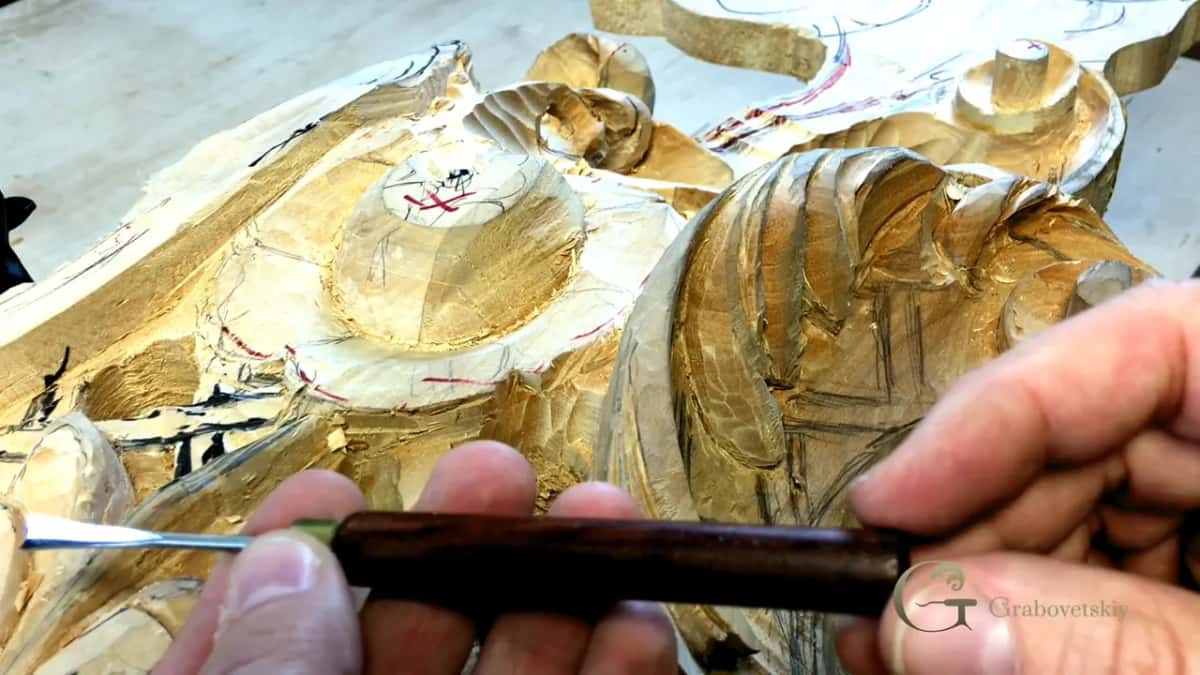Dark bladed tool worked across the gold ground of the carving, handle dark against the leaf.