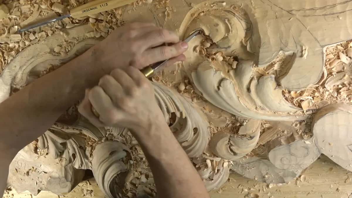More tools lie along the top of the board as two hands lean into a gouge at the hollow.