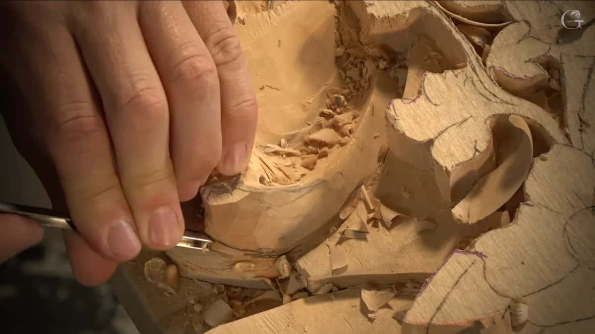 Fingertips guide a small veiner under the edge of the scroll while chips pile in the hollow.