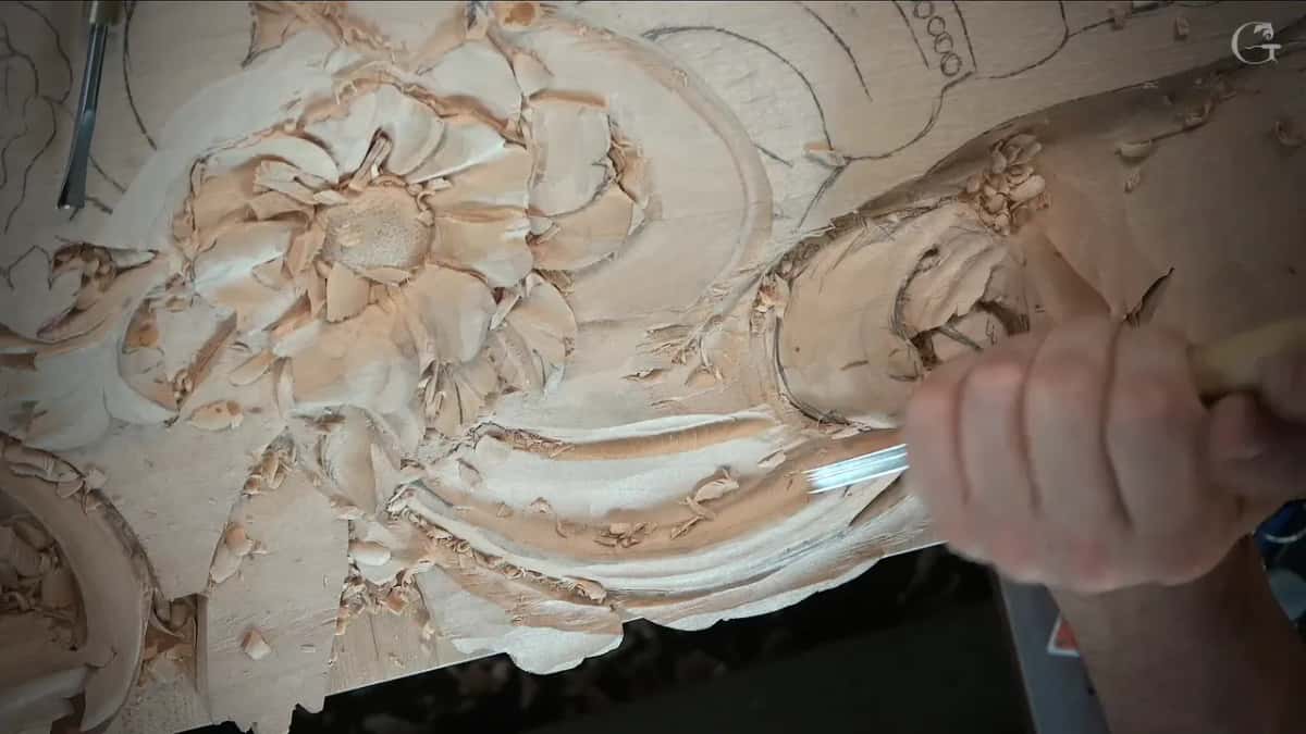 Hands at the scroll again, a pale handled tool going into the hollow beside the flower