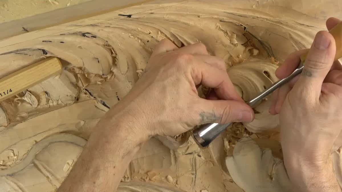 Two hands drive a wide gouge into the waste below the big curl, chips piled around the steel.