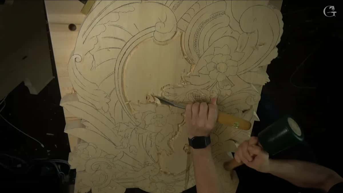 Both hands at work, a gouge in one and a green mallet in the other, above the drawn panel.
