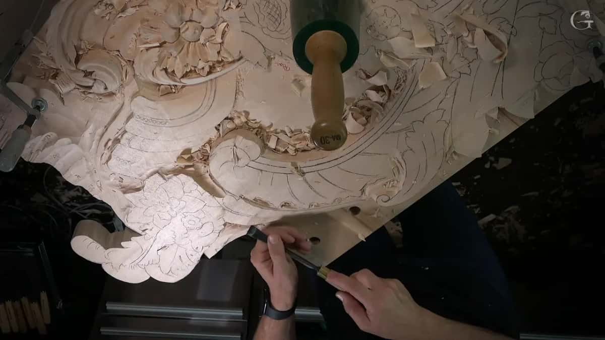 Green mallet resting upright on the carved scroll, both hands down at the bottom corner with a narrow tool
