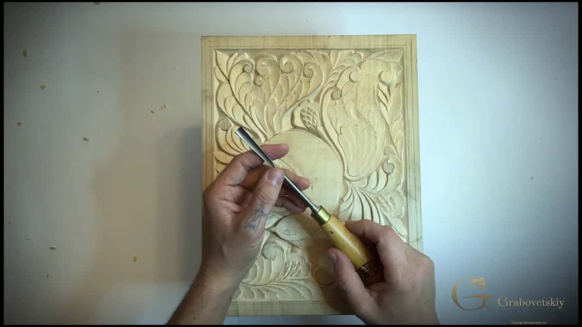 Number 8 gouge held in both hands above the carved panel, brass ferrule and pale wood handle.