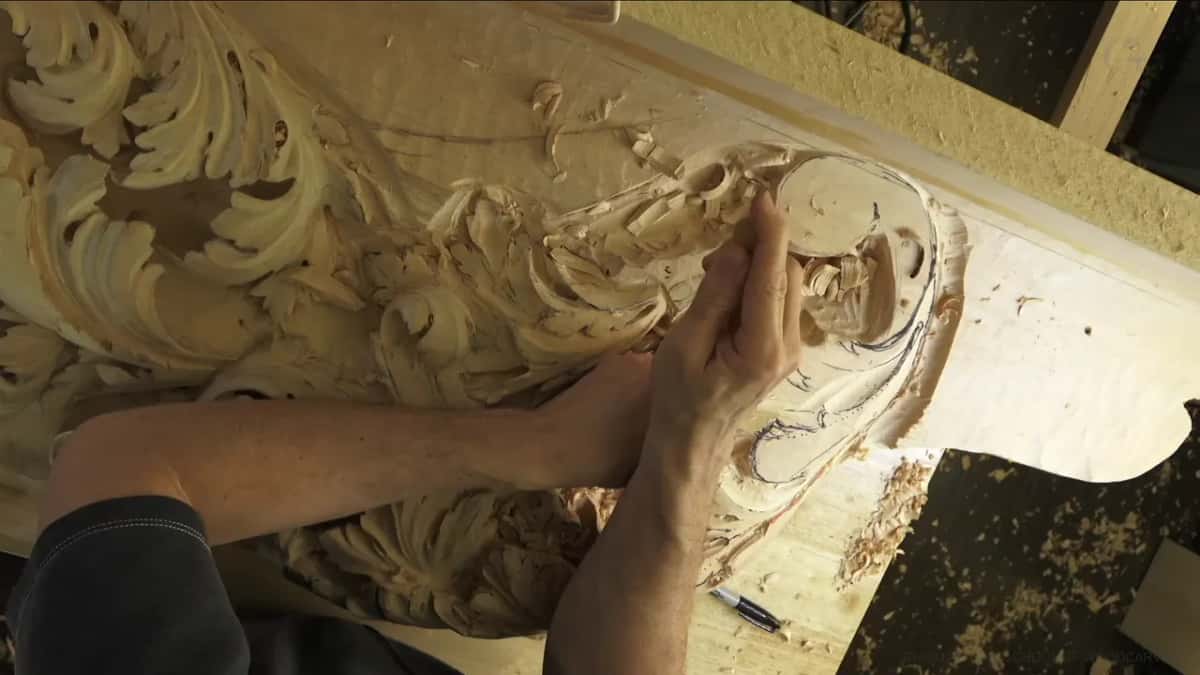 Both hands work near a round boss on the long board, with carved acanthus scrolls off to the left side.