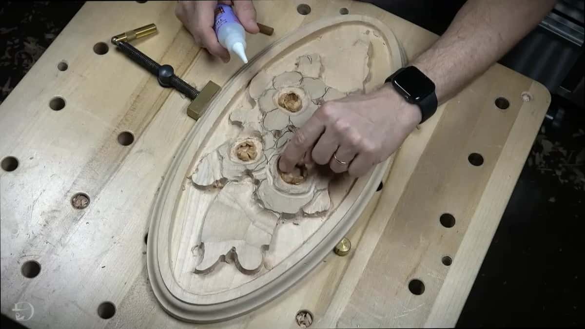 Bottle of thin glue tips over an oval carved plate on the bench while a finger steadies bloom