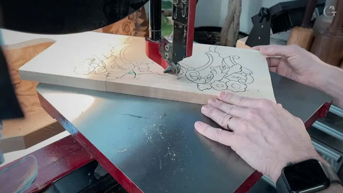 Two hands push a drawn board into a thin band saw blade, scrolls and flowers inked on the wood