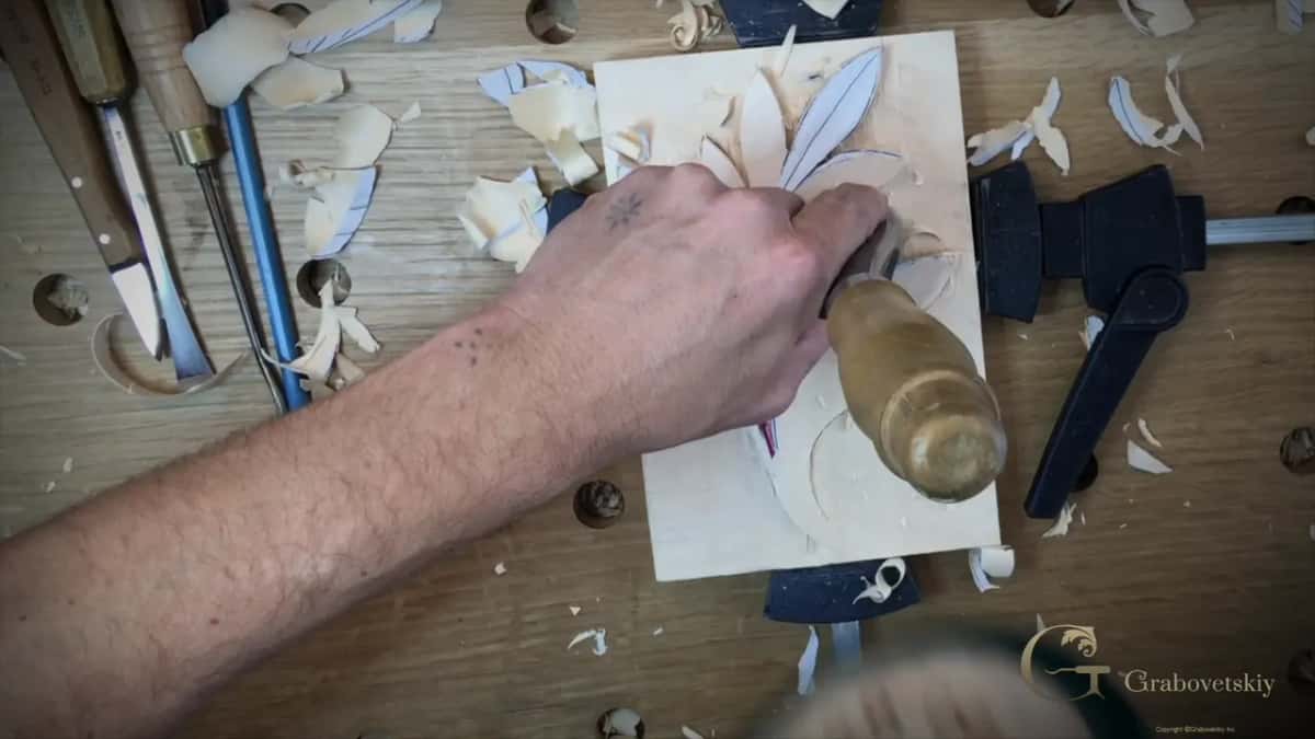 Seen from above, a hand grips a broad gouge with a wood handle on a drawn leaf, board clamped