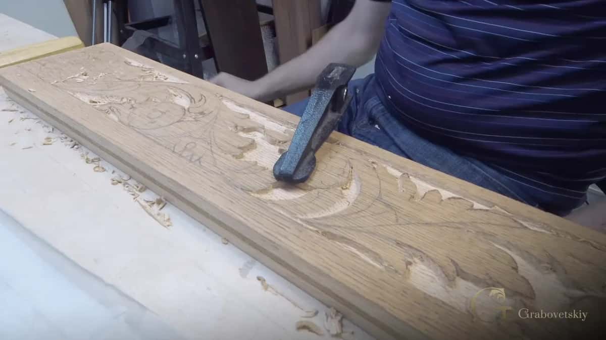 Blue gray mallet head over a long apron board that already shows its carved leaf scrolls