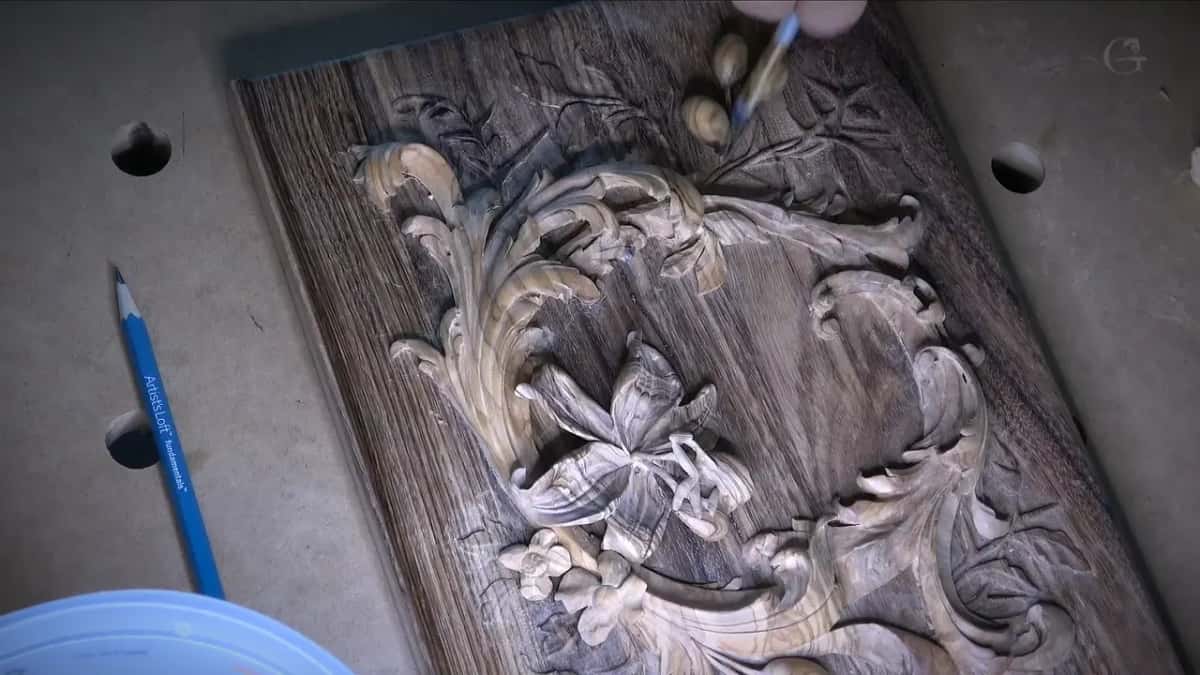 A thin brush touches the carved olives near the top of a dark lid, with a blue pencil lying beside it.