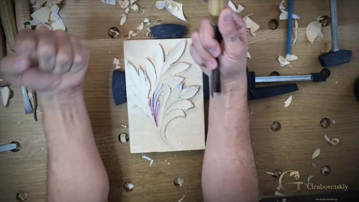 Two forearms raised over the clamped leaf panel, one fist empty and the other gripping a gouge