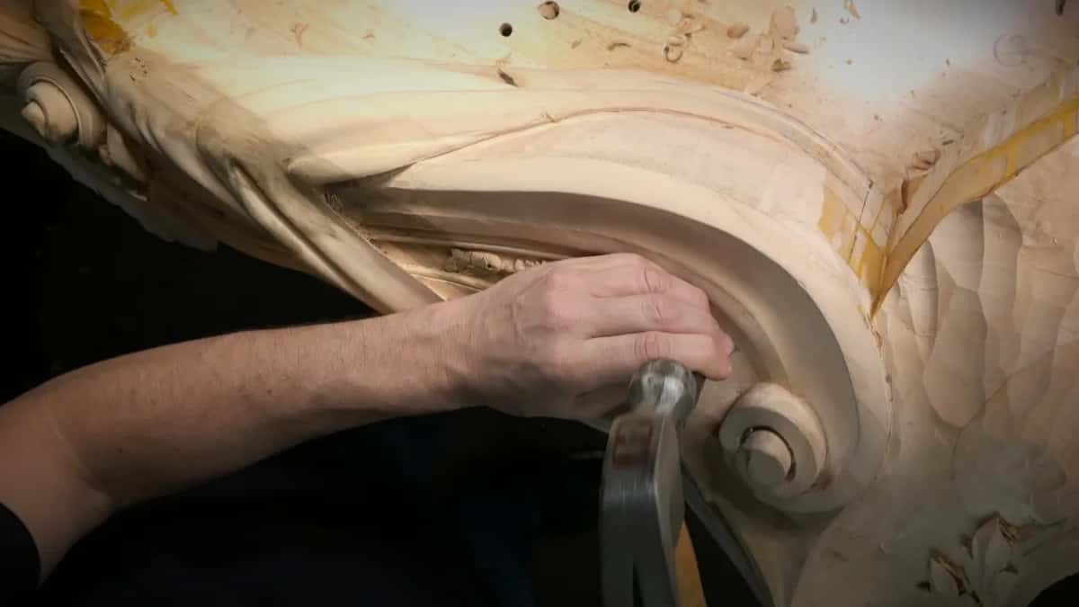 Fingers hold a punch against the ground inside a big carved scroll while a claw hammer waits underneath.