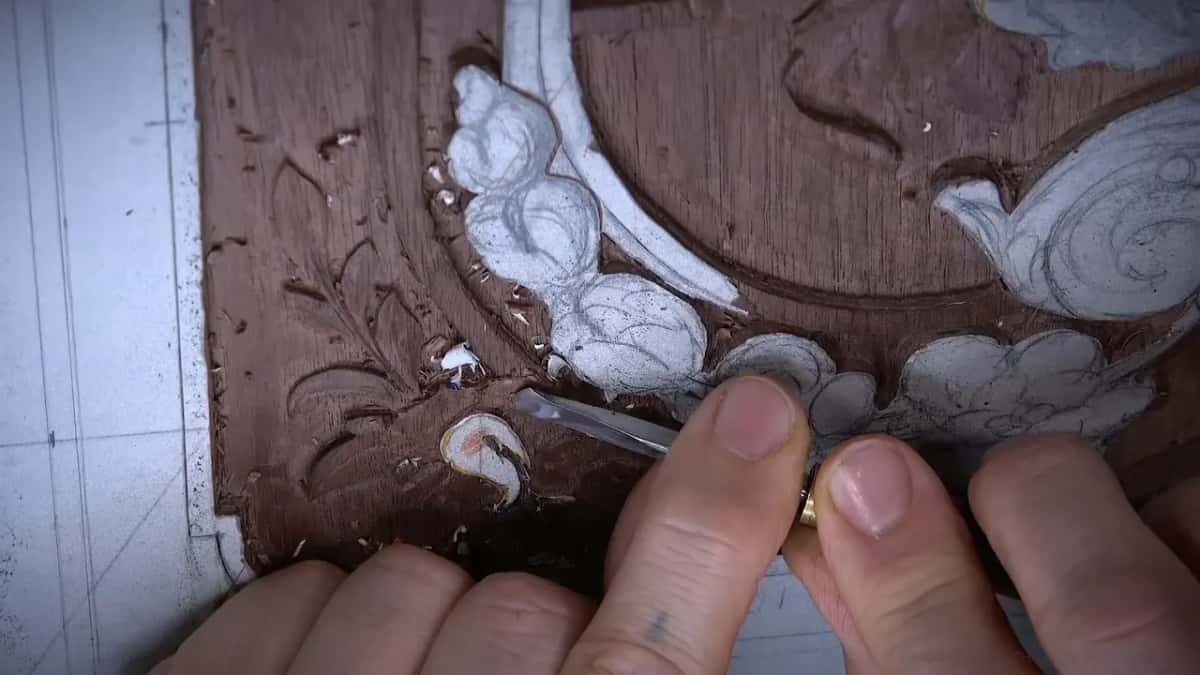 Both hands on a small gouge, its edge in a tight curl just below the garland of flowers