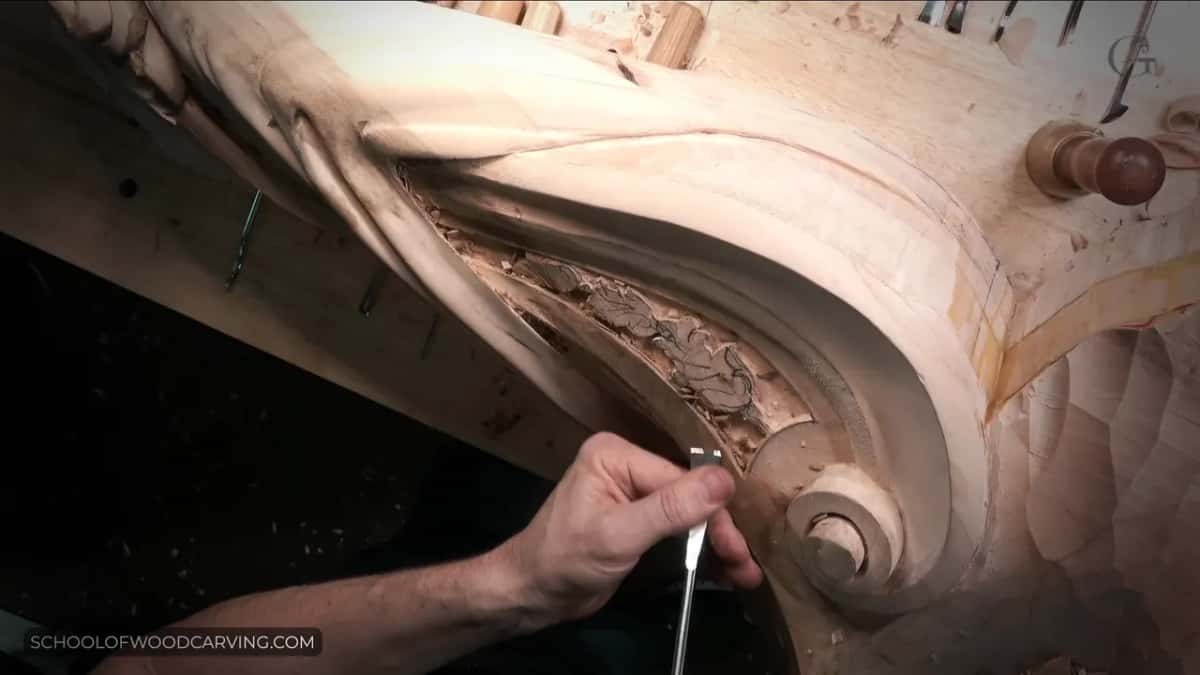One hand guides a narrow chisel beside a rolled volute, with warm light falling over the pale carving.