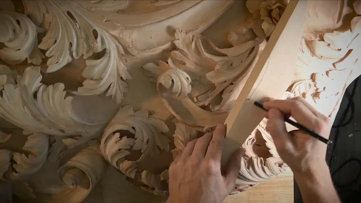 Pencil in one hand and a thin strip of wood in the other, drawing a straight line across acanthus carving