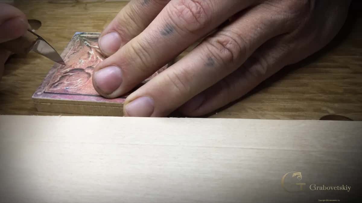 Blade tip going down along a leaf on the reddish paper pattern, my fingers holding the block still.