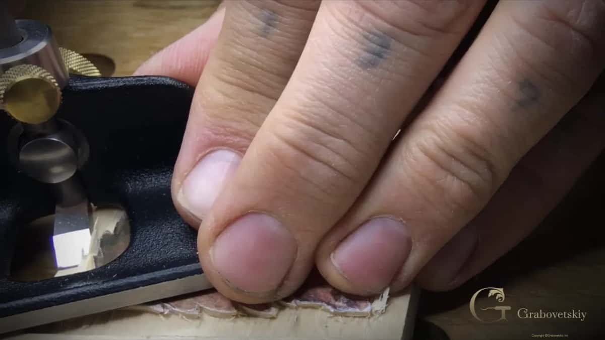 Blade of the router plane seen through its mouth, close to the carving, my fingers pressing the sole flat on the wood.