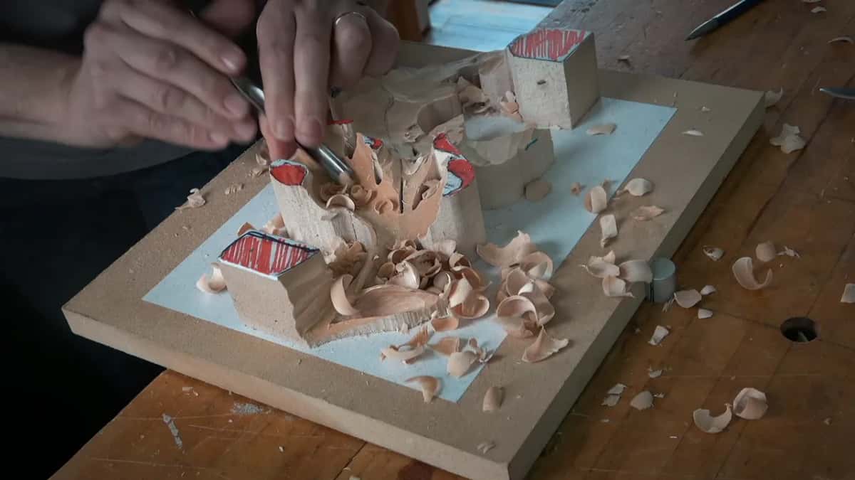 Both hands guide a narrow gouge along the top of the leaf, shavings gathering on the pale blue surface