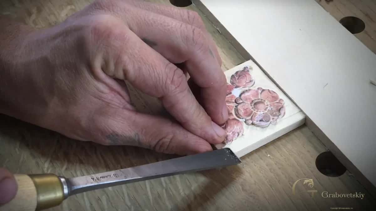 Fingertips pin the block against a white board while a gouge comes in from the lower left