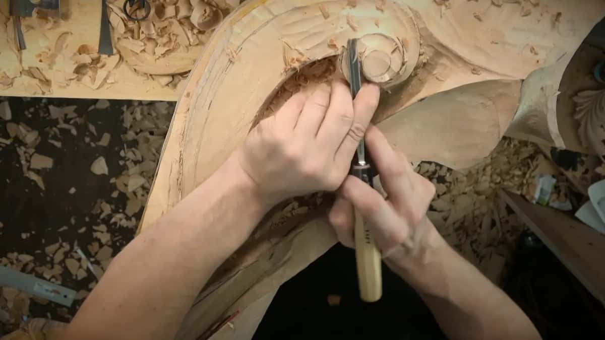 Both hands steady a slim gouge in the hollow beside the center of the scroll, shavings piled over the bench