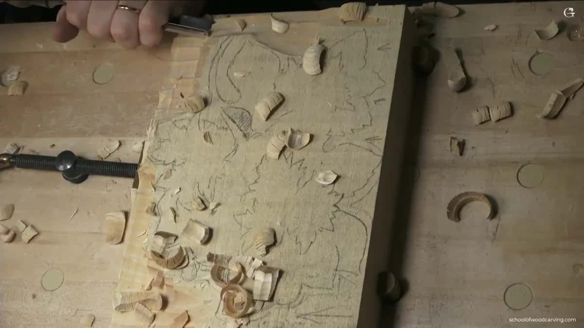 A hand on the gouge at the corner of the traced basswood panel, chips scattered across the top