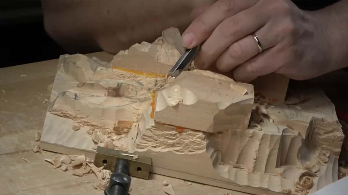Hands steer a small gouge into the tall rough lump just above the brass bench stop