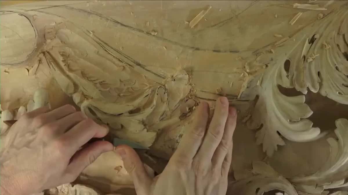 Both hands working a knife along a pencil line, shavings piled up around the scrolls of the carved panel