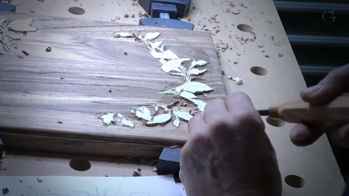 Fingers steady the steel of a light handled gouge as it slices along a pale leaf on the dark board