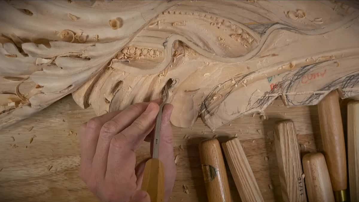 Gouge held low beside a carved eye, a row of handles waiting under it