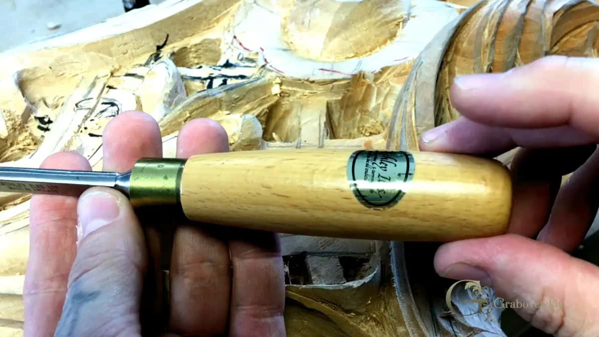 Pale round handle lifted across the fingers, its brass ring and steel pointing left over the carving