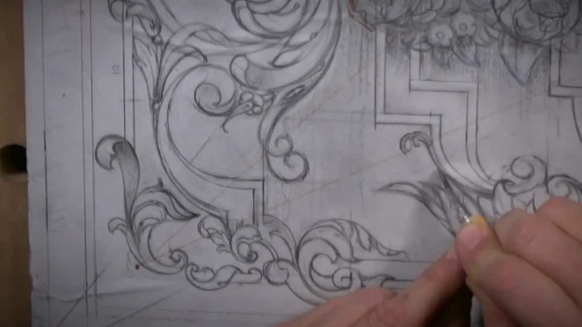Scrolls and leaves drawn in pencil cover the paper, and a hand at the right edge holds a small knife over them.