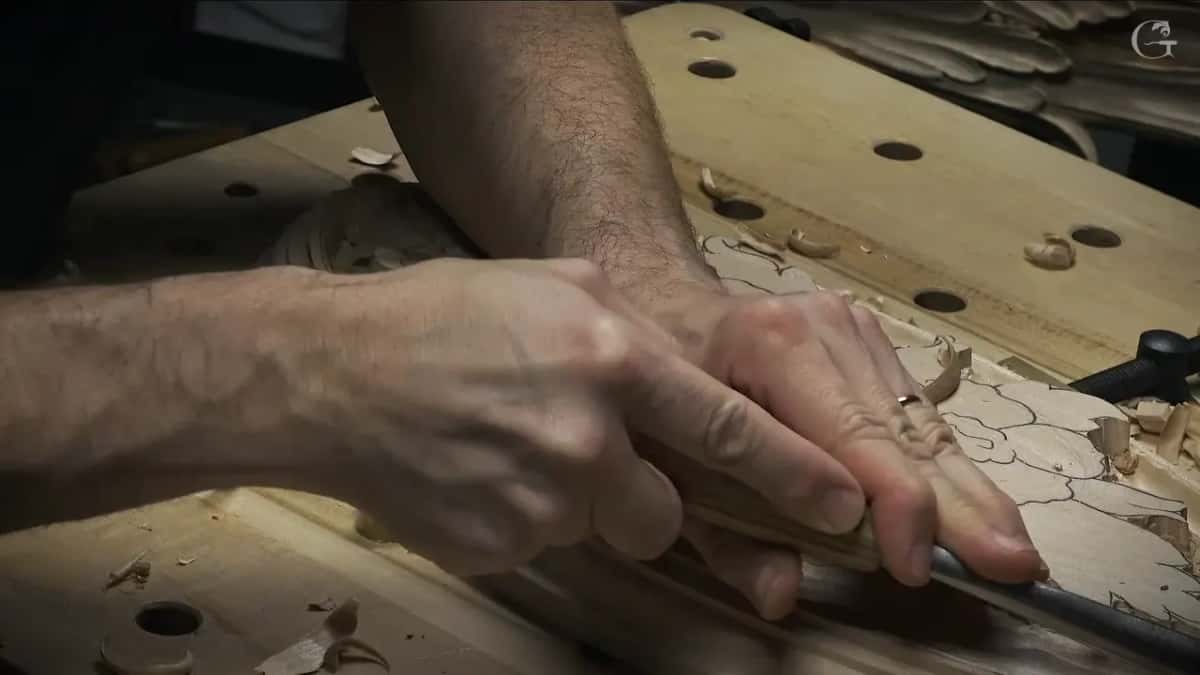 Both hands push a gouge along, lit from one side