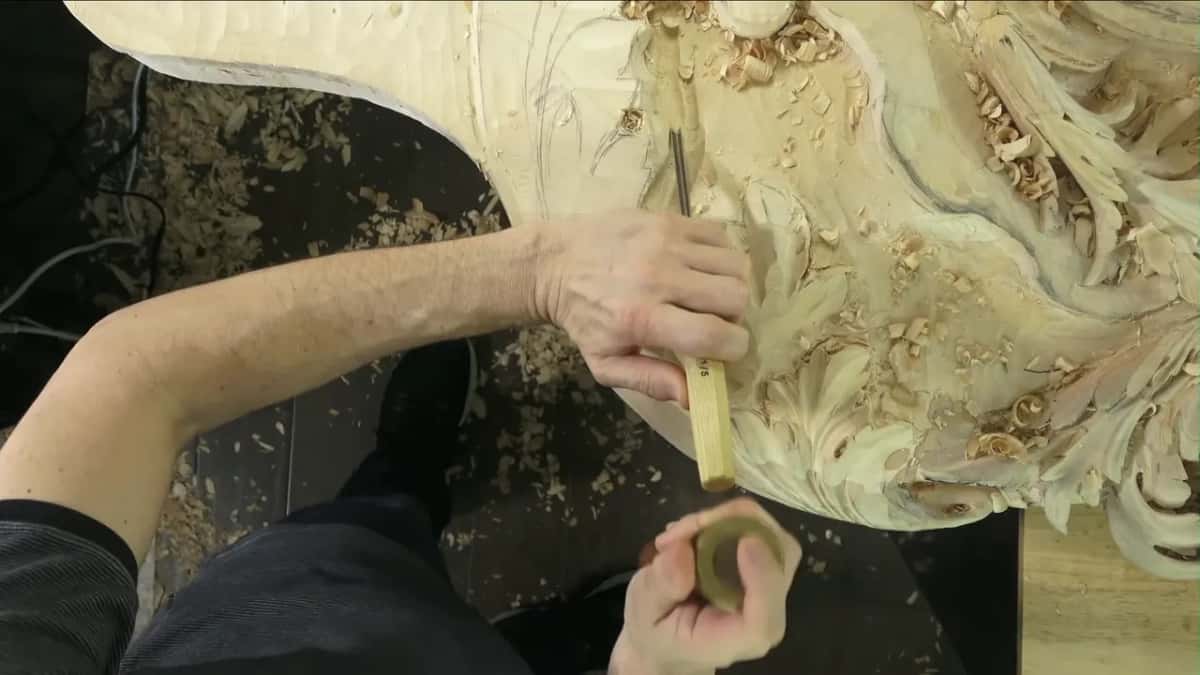 Small brass mallet low in one hand, the other setting a gouge against the carved acanthus overhead