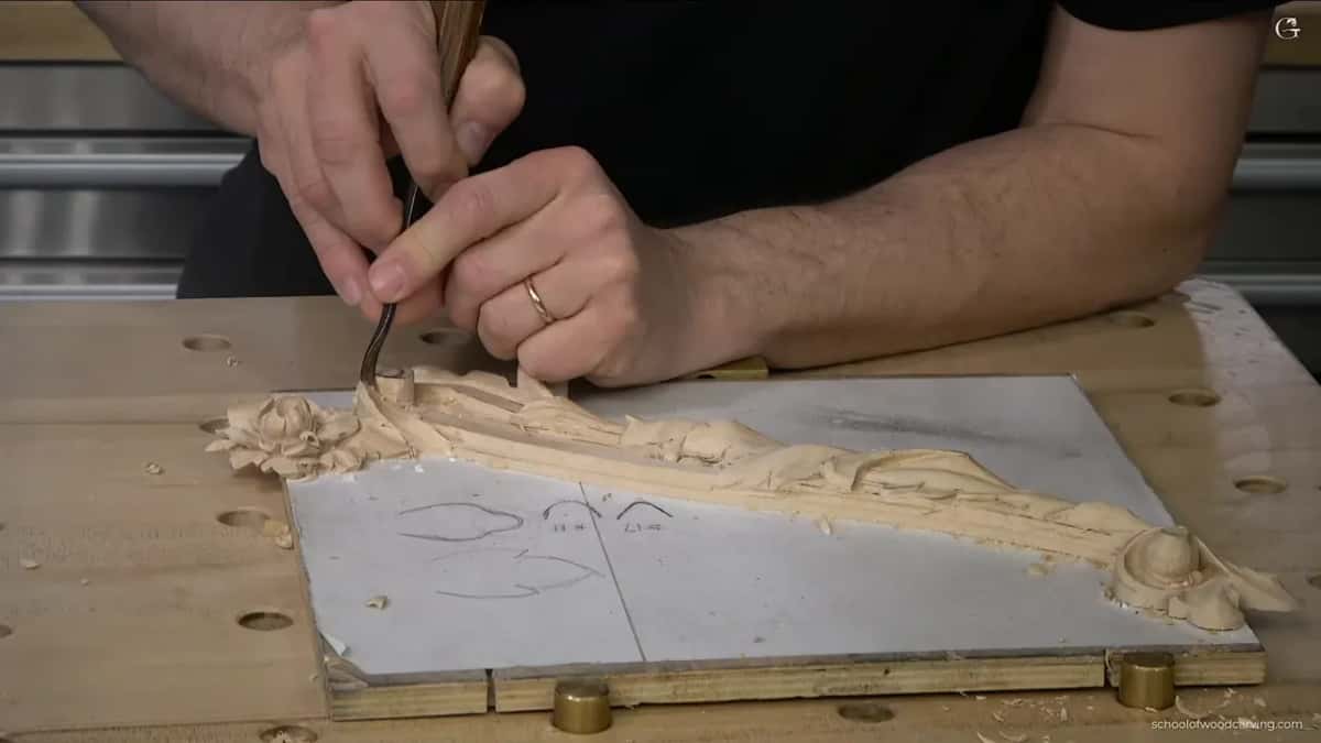 Both hands guide a gouge along a carved molding, a flower at its far end