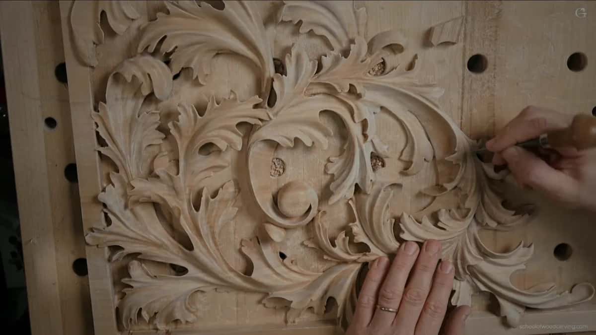 A carving knife held low scrapes a curling acanthus leaf while the other hand rests flat on the panel.