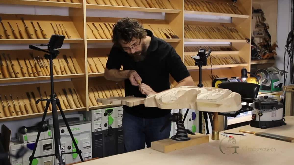 A carver bends over a big scroll on a stand, with tool racks behind
