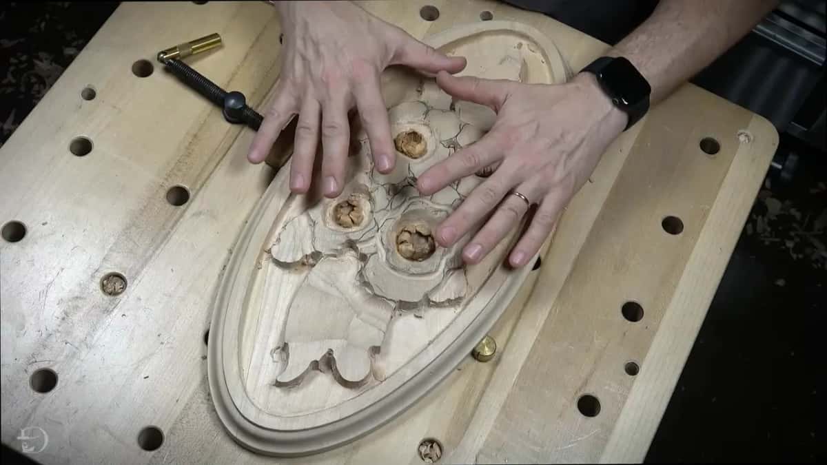 Two hands rest on an oval rosette carving with four small flowers, held on a bench full of holes