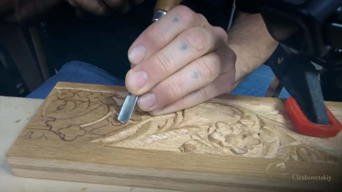 Hand holding a V tool at the pencil outline of a small flower, carved flowers further along