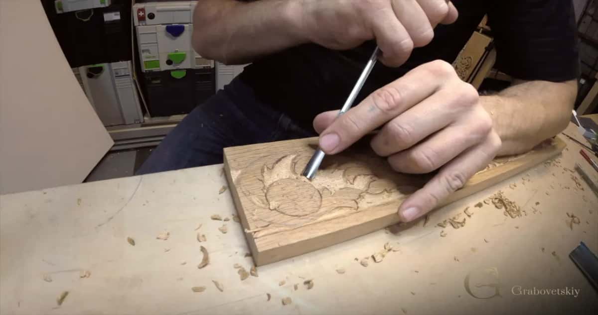 Both hands guide a gouge around a flower ring near the end of the board, chips scattered across the bench